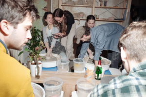 People are laughing and drinking while they play on pottery wheels.